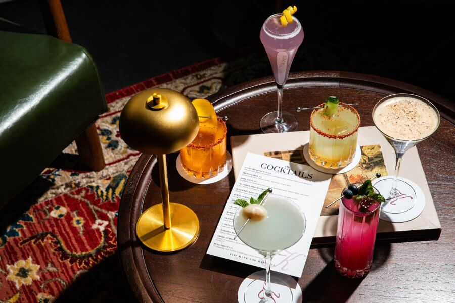 A selection of colorful cocktails graces a dark wooden table alongside a gold lamp and menu, set atop a patterned rug. This inviting scene is the perfect snapshot of one of Houston's hottest new restaurant experiences.