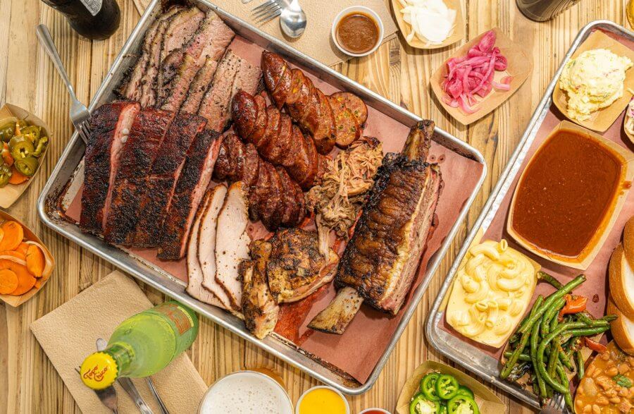 A metal tray loaded with grilled meats—ribs and sausage—accompanied by sides like macaroni, pickles, and beans on a wooden table. Nearby, drinks and condiments complete this feast.
