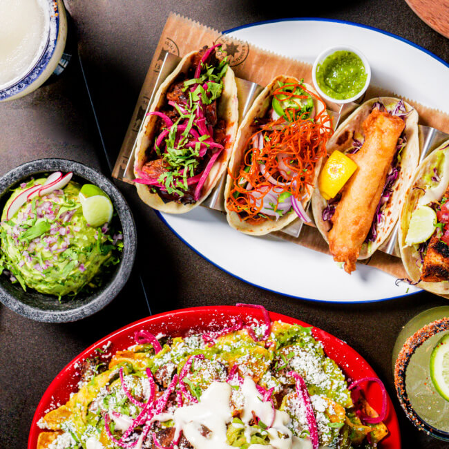 9 New Nashville Restaurants to Try This Month - 20 A plate of assorted tacos with various fillings, a bowl of guacamole with lime, and loaded nachos topped with cheese and sauce.
