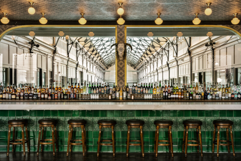 9 New Nashville Restaurants to Try This Month - 28 A bar with green tile and seven empty barstools stands in front of a large mirror reflecting a bright, glass-roofed arcade—a stylish setting typical of new Nashville restaurants. Bottles are neatly arranged on the countertop.