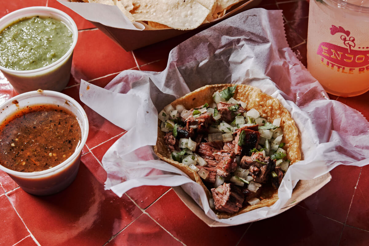9 New Nashville Restaurants to Try This Month - 23 Indulge in a beef taco topped with diced onions and herbs, perfectly wrapped and served with red and green sauces alongside a refreshing pink drink. Discover this culinary delight on a vibrant red tiled surface.