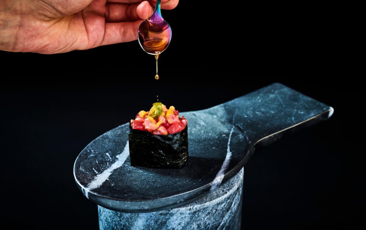 In March 2025, a hand delicately drizzles sauce from a spoon onto a gourmet sushi piece topped with diced ingredients, artfully placed on a stone serving dish against a black background—perhaps a culinary masterpiece found in one of Nashville's new restaurants.
