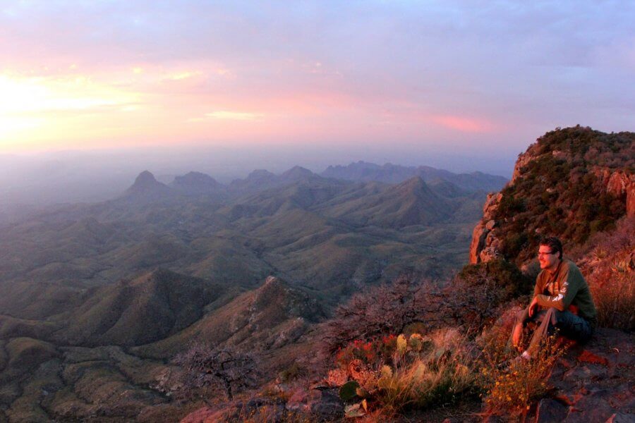 Person sitting on a rocky ledge, overlooking a vast Texas landscape of mountains and valleys at sunset, with a mix of warm and cool tones in the sky, capturing the essence of serene Texas hikes.