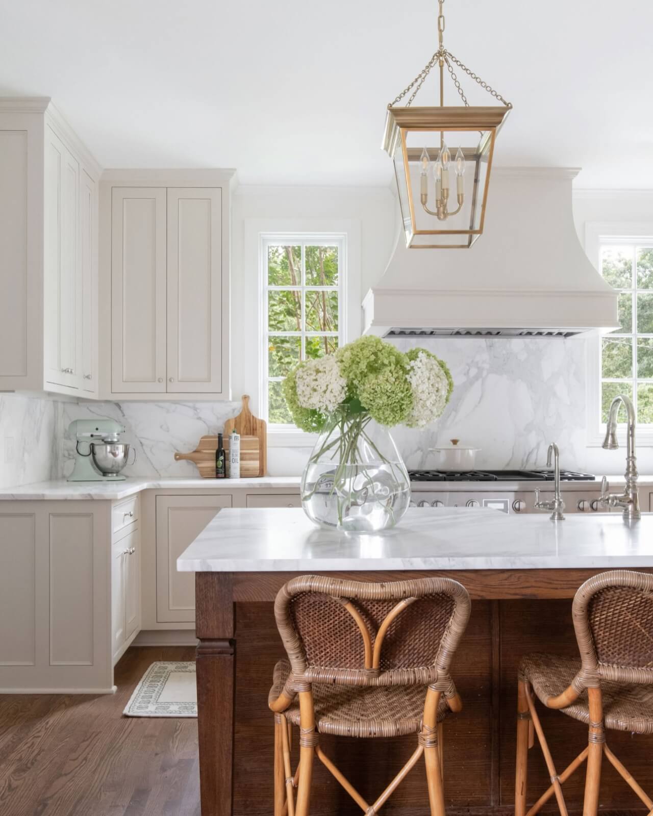 Bright kitchen with white cabinets, marble countertops, a large vase of hydrangeas on the island, wicker barstools, and gold pendant lighting—an inspiring design often seen from the Best Interior Designers in Nashville.