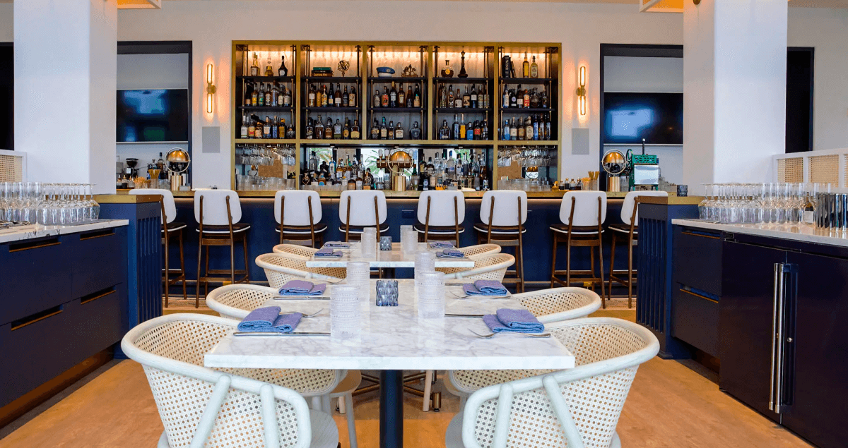 Sip and savor at this modern bar with a marble counter and wicker chairs, surrounded by tables. Enjoy a well-stocked backlit shelf of bottles during your 30A Happy Hour Tour. Napkins are neatly folded on each table, ensuring an inviting atmosphere.
