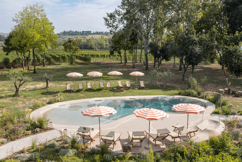 Nestled in a tranquil garden setting, Villa Yeppa offers a luxurious outdoor pool adorned with lounge chairs and umbrellas. With trees and the Italian countryside as your backdrop under a partly cloudy sky, it's the ultimate retreat.