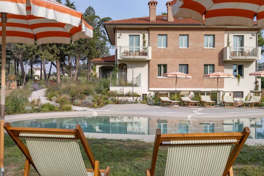 Two lounge chairs face a kidney-shaped pool beside Villa Yeppa. Orange and white striped umbrellas provide shade. The villa, with its brick exterior and multiple windows, offers a luxury retreat in Northern Italy, with trees gently swaying in the background.