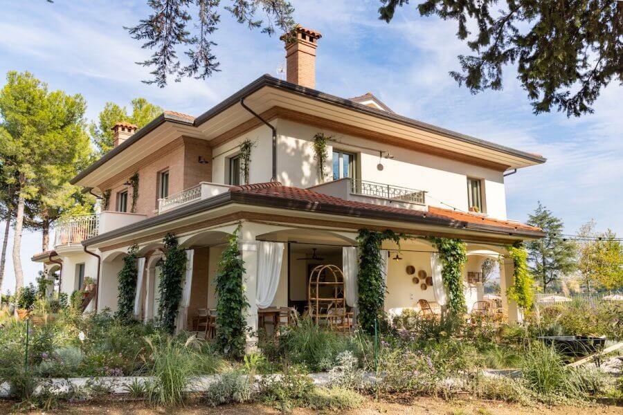 This luxury Italian retreat, Villa Yeppa, is a two-story house with beige walls and brick accents, nestled in the lush greenery of Northern Italy. It features balconies, a covered porch with curtains, and outdoor seating for a truly enchanting experience.