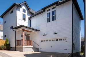 A charming two-story white house on Moore Ave in Nashville, TN, features a wooden porch and double garage door. The concrete driveway complements the black roof and window trims. A clear blue sky serves as the perfect backdrop for this picturesque home.