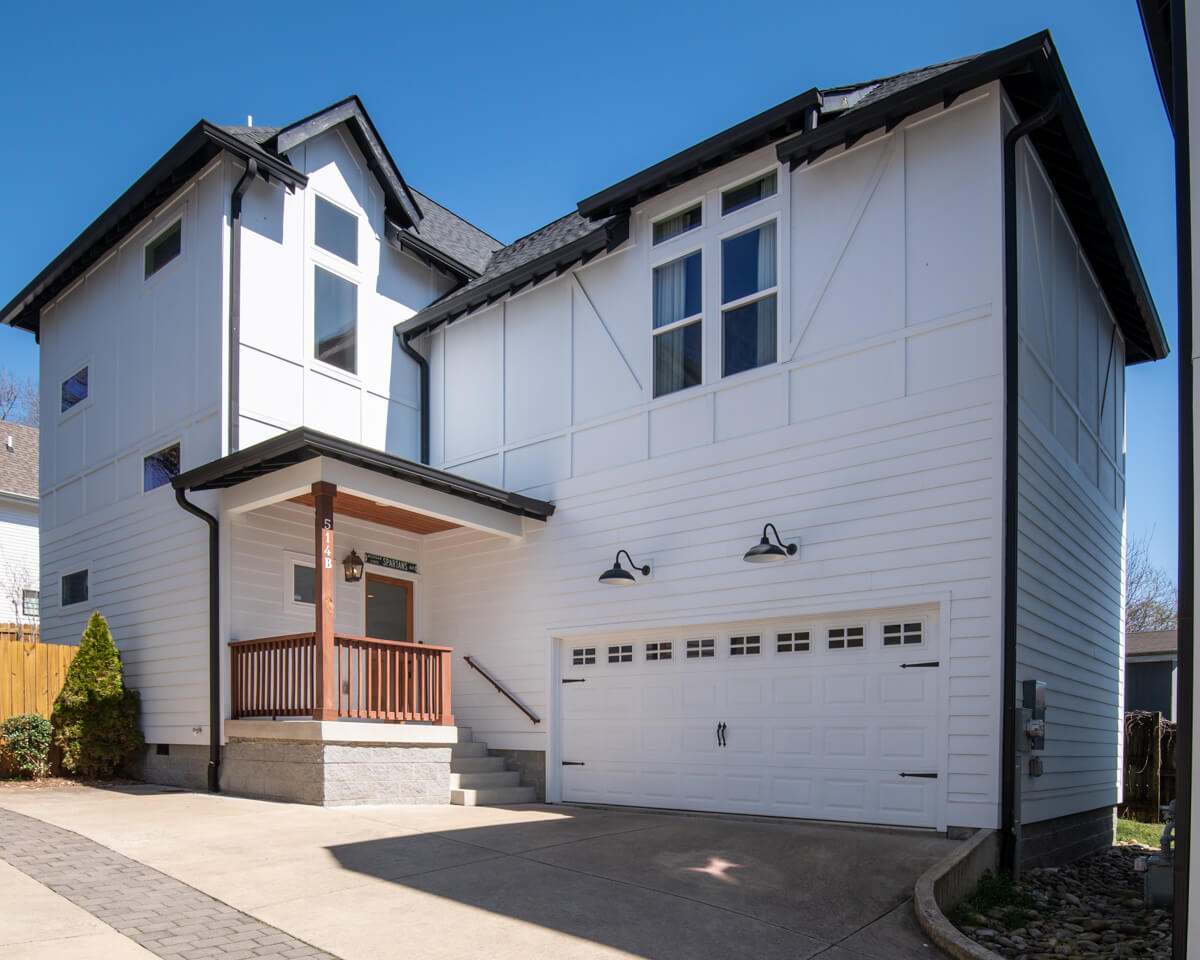 A charming two-story white house on Moore Ave in Nashville, TN, features a wooden porch and double garage door. The concrete driveway complements the black roof and window trims. A clear blue sky serves as the perfect backdrop for this picturesque home.