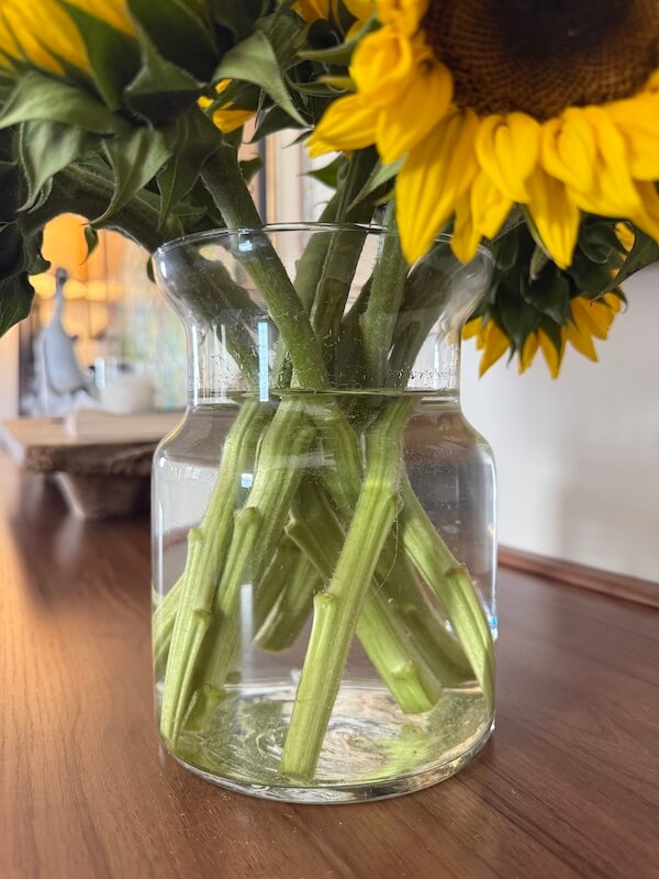 A vibrant floral arrangement of sunflower stems in a glass vase with water sits on a wooden table, brightening any room.