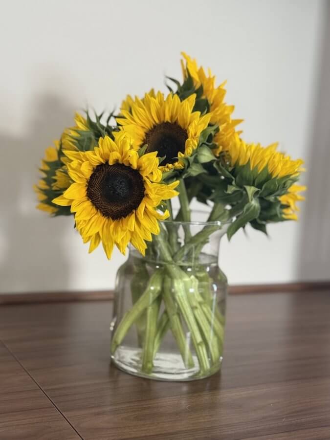 A floral arrangement in a glass vase holds yellow sunflowers with green stems, brightening the wooden surface.