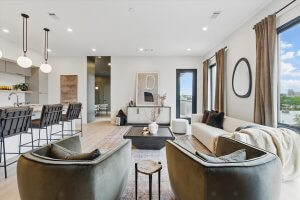 Modern open-concept living room at 500 Merritt Ave, Nashville TN, featuring neutral decor, two green armchairs, a white sofa, large windows, and a kitchen area with barstools and pendant lights in the background.