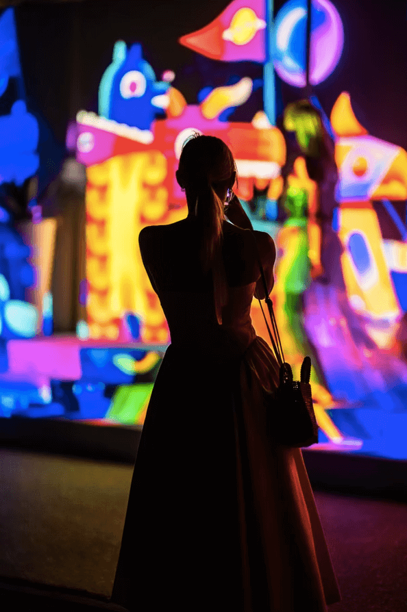 A person in a dress stands facing a brightly colored, abstract neon art display, reminiscent of Digital Graffiti at Alys Beach—an illuminated escape into vibrant creativity.