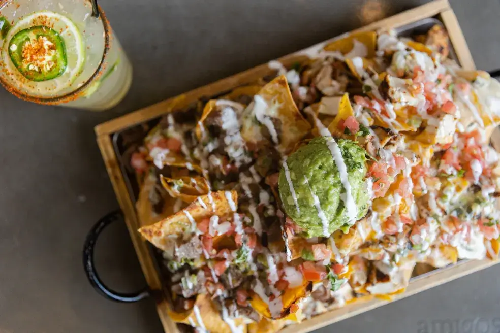 A tray of loaded nachos topped with guacamole, diced tomatoes, and melted cheese sits next to an iced drink with lime and jalapeño—perfect for enjoying elevated Mexican dining on 30A.