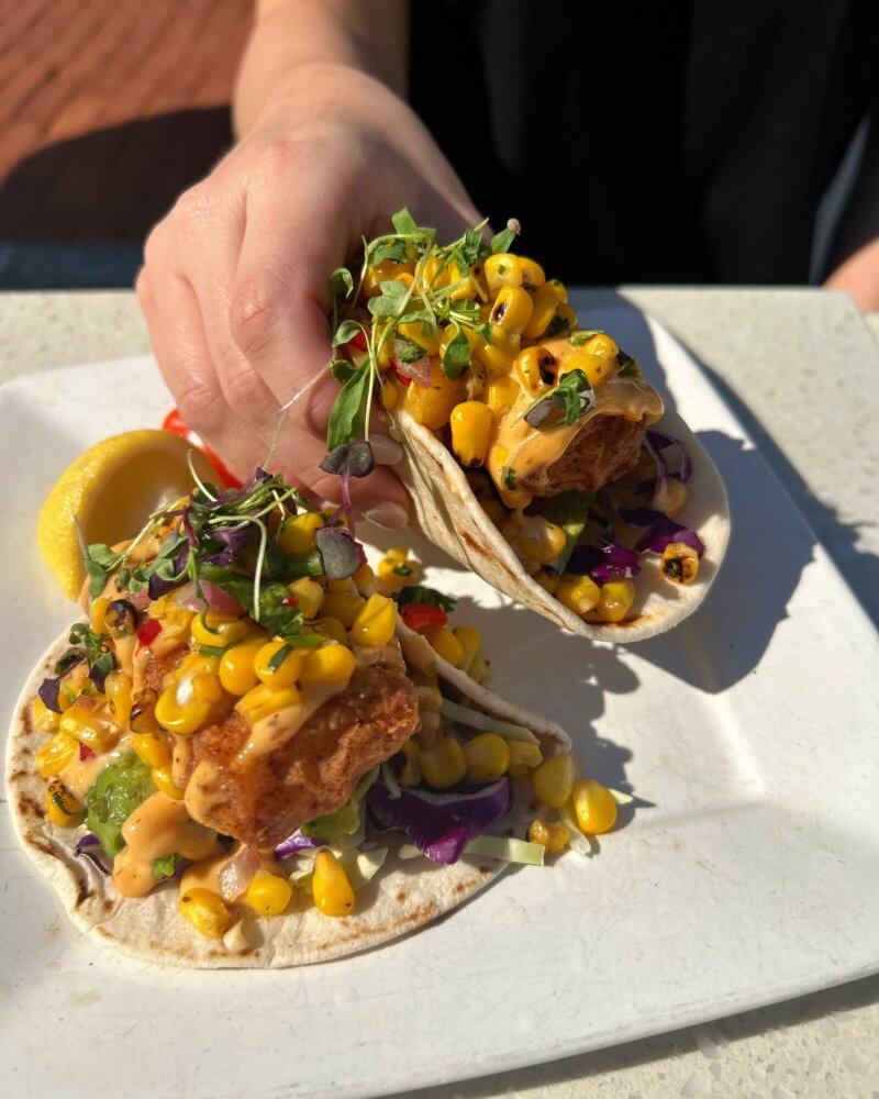 A hand holds two street-style tacos filled with fried fish, corn, microgreens, shredded cabbage, and sauce—an elevated Mexican dining experience on a white plate with a lemon wedge, perfect for savoring the flavors of 30A.