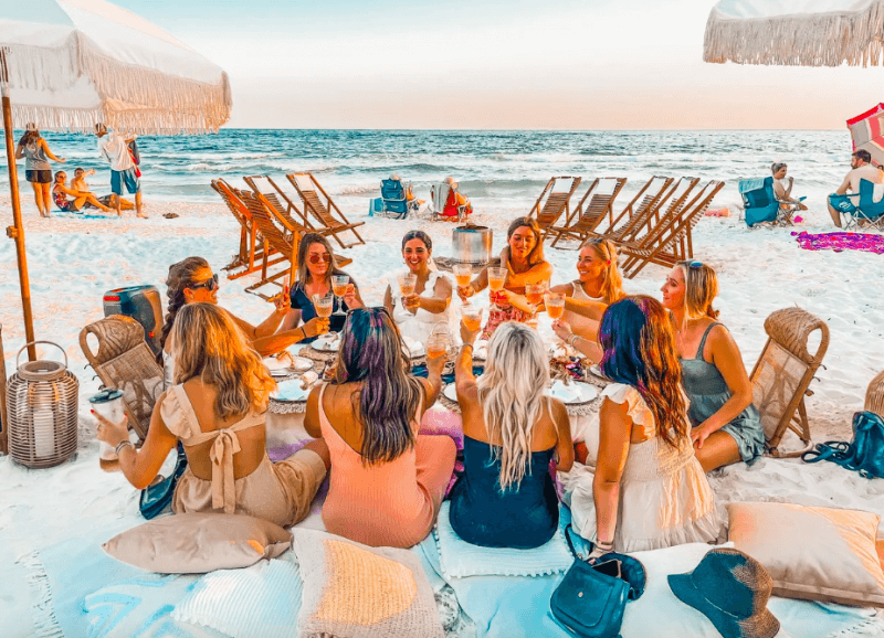 A group of people sits on a picturesque 30A beach, holding glasses and toasting. They're surrounded by pillows and a low table laden with food, near the ocean. Wooden lounge chairs and umbrellas dot the landscape, promising unique adventures in this coastal paradise.