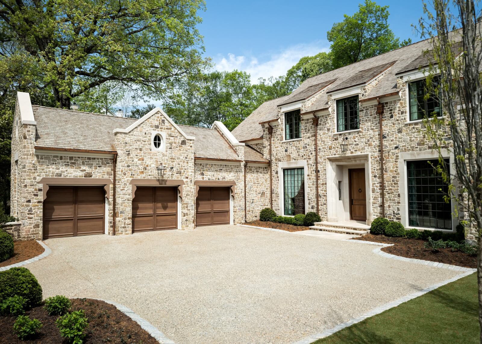 A stone-faced two-story house with three brown garage doors, large windows, and a gravel driveway, surrounded by manicured shrubs and trees.