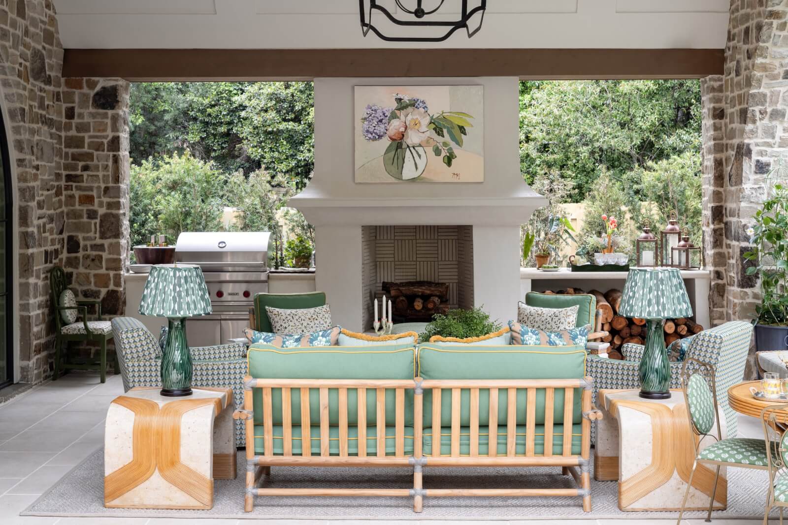 An outdoor living area inspired by the English Countryside, with green cushioned furniture facing a fireplace, flanked by lamps and tables, a grill on the side, and lush greenery.