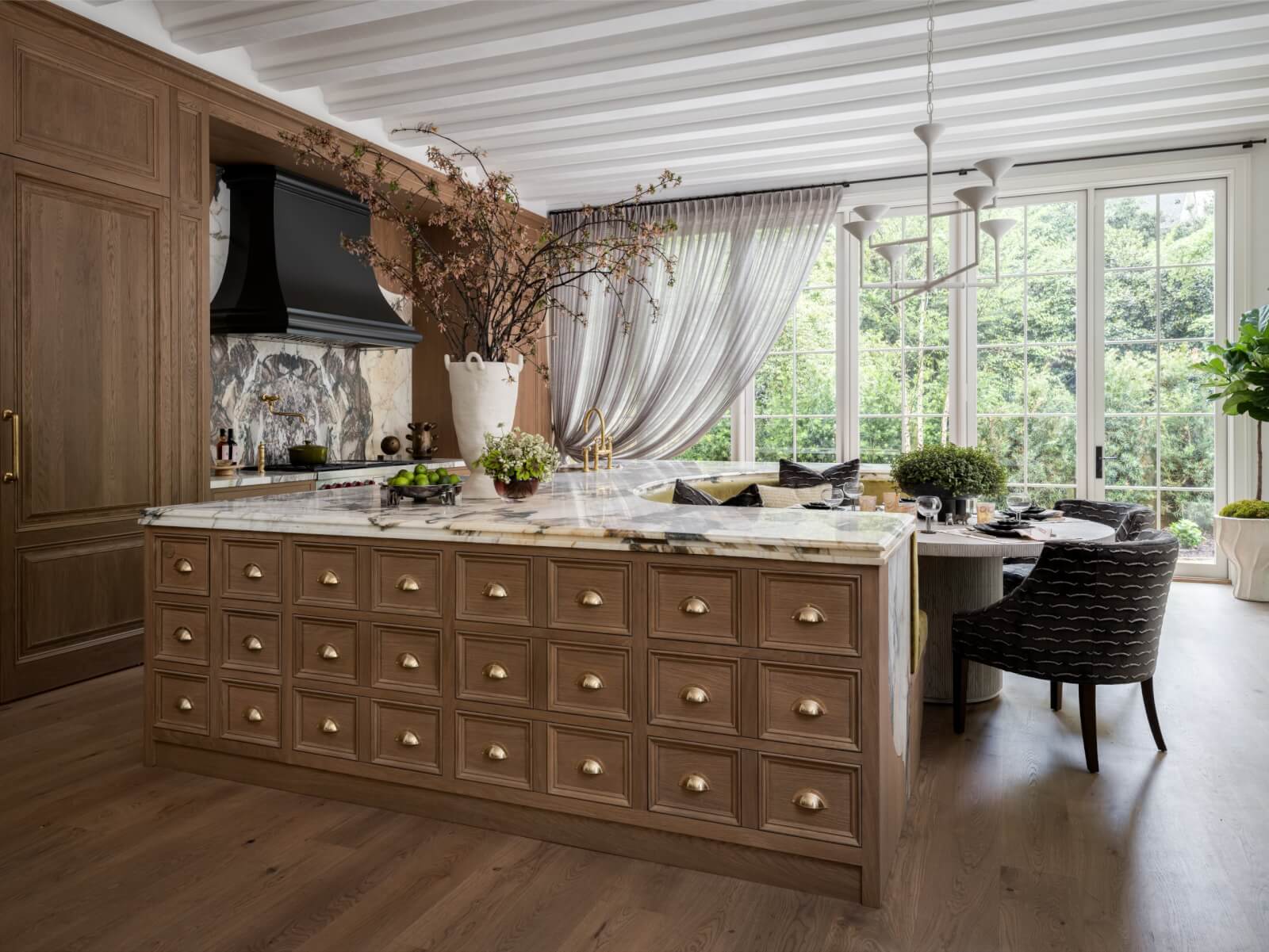 A modern kitchen inspired by the English Countryside, with a large marble island featuring many drawers, wood cabinets, a dining area, and tall windows offering views of greenery.