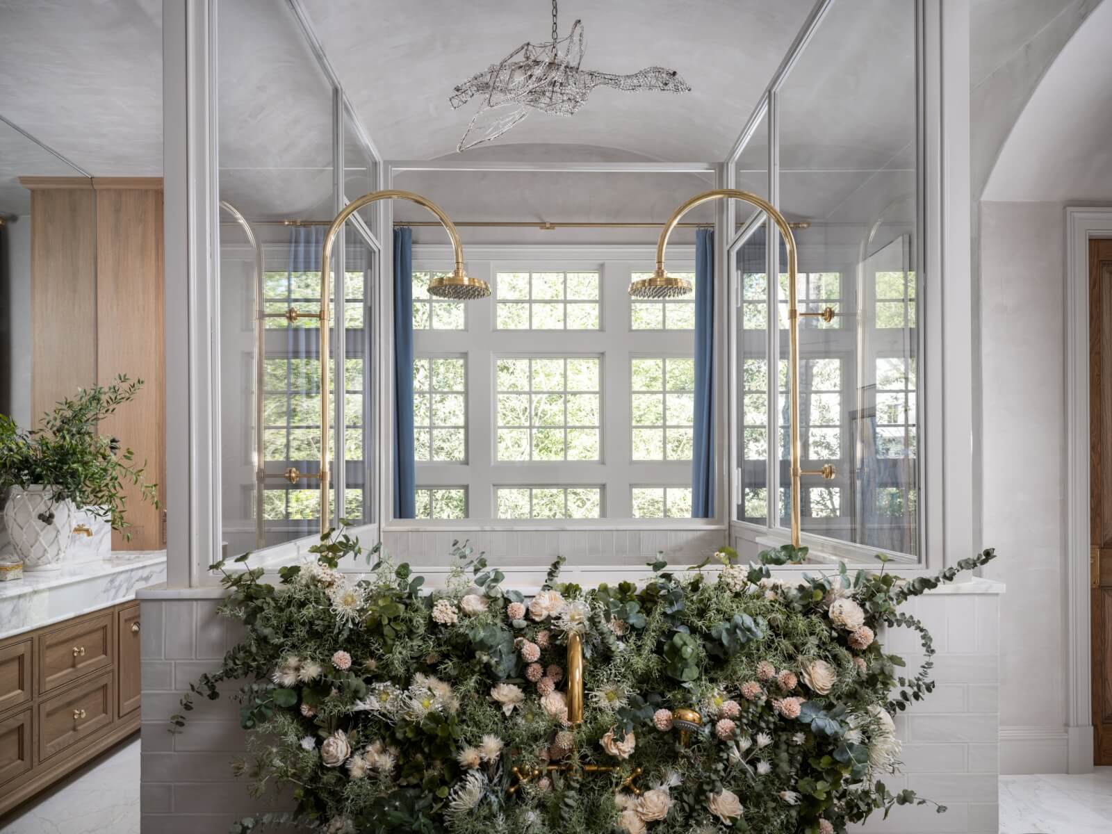 Modern bathroom inspired by the English Countryside, featuring a glass shower enclosure, dual gold showerheads, large grid windows, and a tub adorned with lush greenery