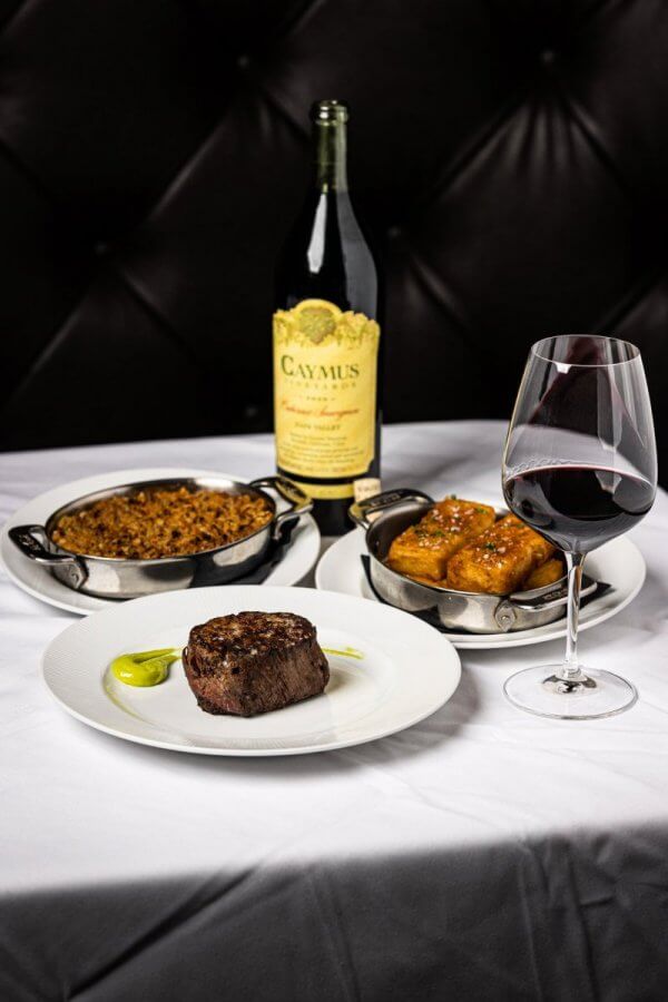 Steak with sauce, a bottle of red wine, a glass of wine, a dish of rice, and a dish of bread rolls on a white tablecloth create unforgettable dining experiences. 