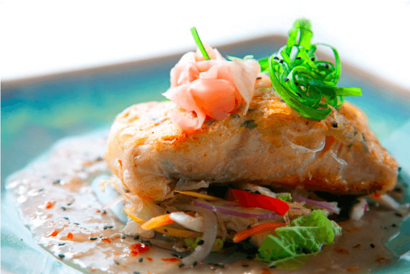 Enjoy an elevated dining experience with a grilled salmon fillet on vibrant vegetables, draped in a creamy sauce. Garnished with pink pickled ginger and green seaweed, it's elegantly presented on a blue plate.