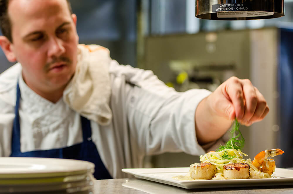 In the kitchen, a chef masterfully garnishes a plated dish with scallops and greens, creating an elevated dining experience.