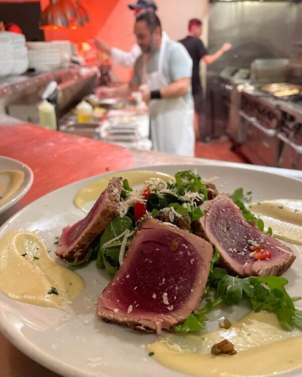 In a bustling 30A dining atmosphere, a plate of seared tuna rests on a bed of arugula, topped with grated cheese and sauce. Chefs work in the background, crafting elevated dining experiences that are well worth booking ahead for.