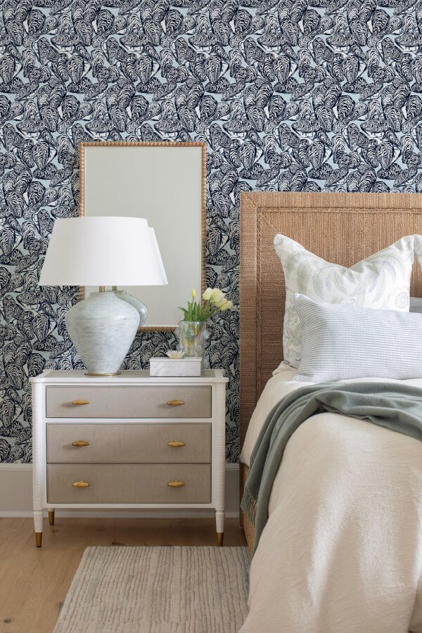 Bedroom scene with a bed, white nightstand, lamp, and decorative items. The wallpaper has a navy blue botanical pattern. The bed is neatly made with a light beige blanket and several pillows.