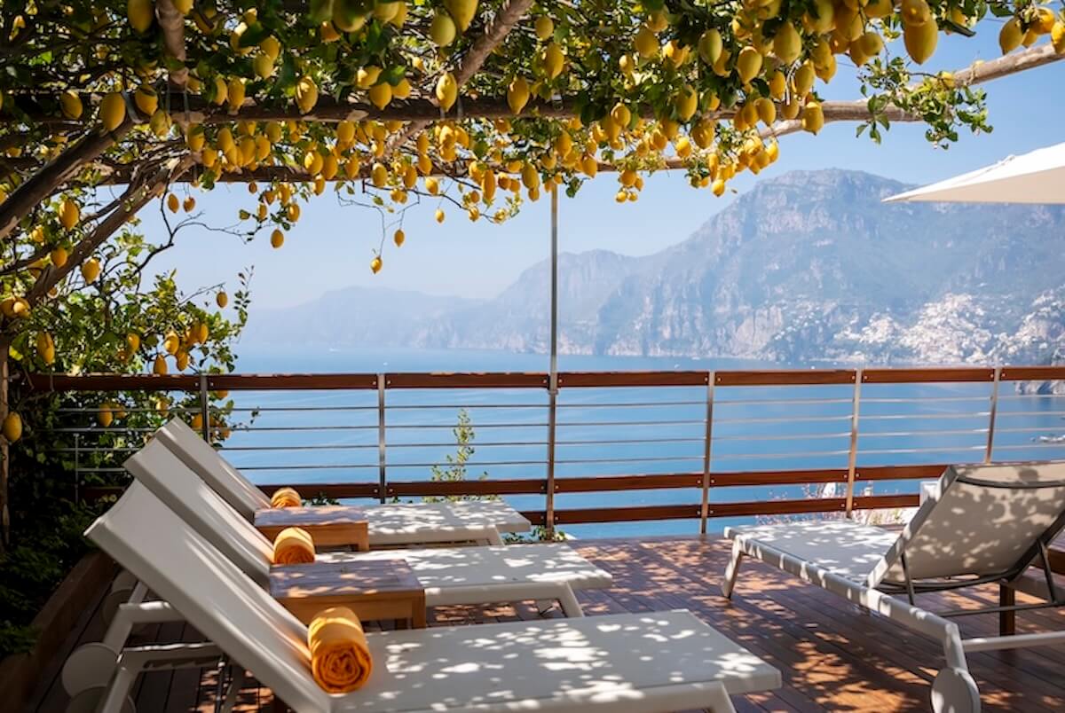 Four luxury lounge chairs with orange towels rest on a wooden deck under a lemon tree. Overlooking a railing, the hidden gem offers breathtaking views of distant mountains and the sea on the Amalfi Coast. A white umbrella provides soothing shade.
