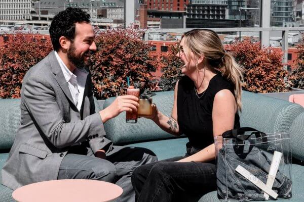 On the stylish rooftop patio of Thompson Nashville, two people raise their glasses in a cheerful toast. Seated on a blue couch, the man sports a gray suit while the woman, elegantly dressed in black with a transparent bag by her side, enjoys the view of red bushes and city buildings.