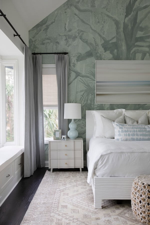 A bedroom with a white bed, nightstand, and lamp. The wall features a tree mural. A window with grey curtains and a bench is beside the bed. A patterned rug covers the dark floor.