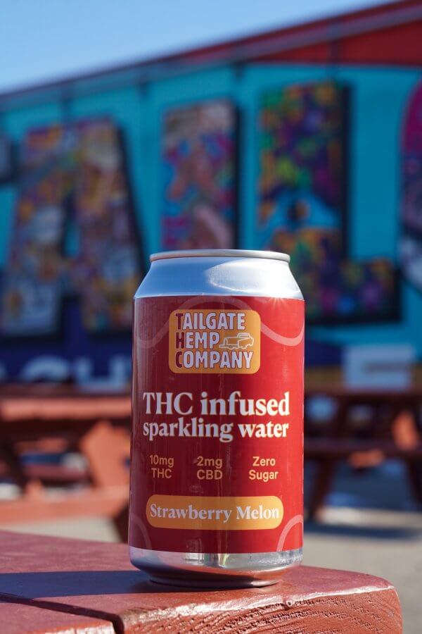 A can of Tailgate Hemp Company THC infused sparkling water in strawberry melon flavor sits on a picnic table with a colorful mural in the background, celebrating the fun spirit of TailGate Brewery.