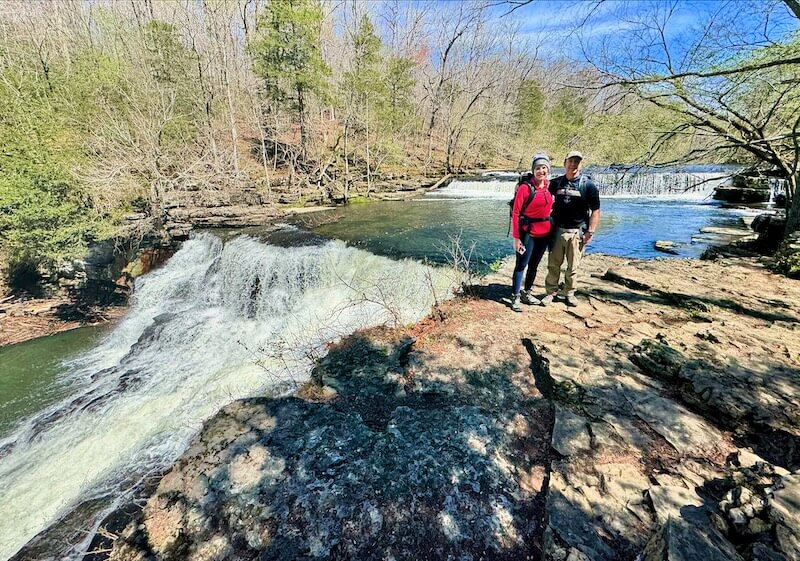 Two people stand on a rocky ledge near a flowing waterfall, surrounded by trees and a clear blue sky, enjoying their 3-mile waterfall hike just outside of Nashville.