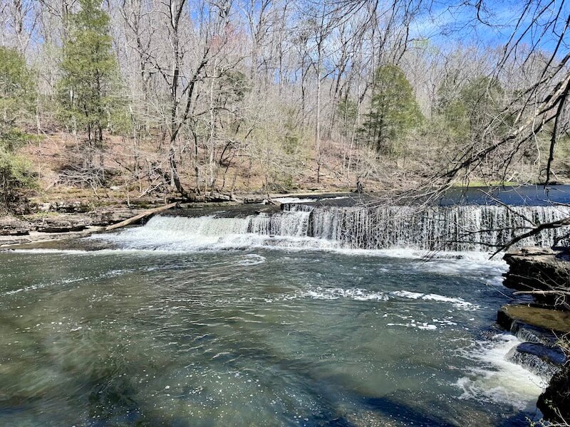 A small waterfall flows over a rocky ledge surrounded by leafless trees and some greenery, under a clear blue sky, creating the perfect backdrop for a serene 3-mile waterfall hike near Nashville.