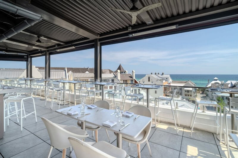 Experience elevated dining at a rooftop restaurant, where sleek white tables and chairs provide the perfect setting. Overlooking rooftops with a stunning ocean view under a clear blue sky, it's the ideal spot along 30A. 