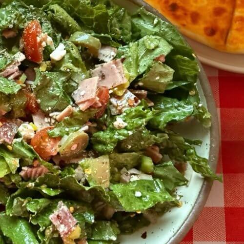 A delicious salad with leafy greens, chopped tomatoes, sliced meat, and cheese sits elegantly on a plate. Hinting at rustic charm, part of a round bread rests nearby on the classic red and white checkered tablecloth—your perfect recipe for a refreshing meal.