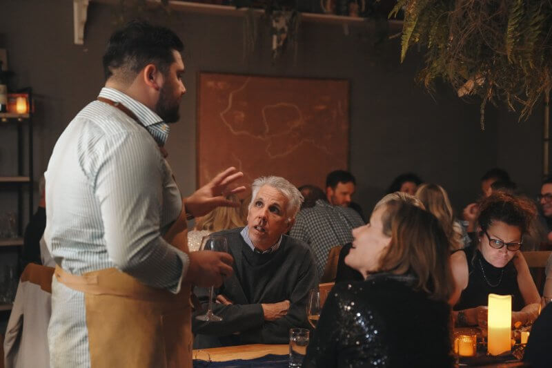 In a dimly lit restaurant known for its elevated dining experiences, a man in an apron speaks to three seated patrons. While one checks their phone, the others listen attentively, savoring the insider tips on 30A's hidden gems and the importance of booking ahead.