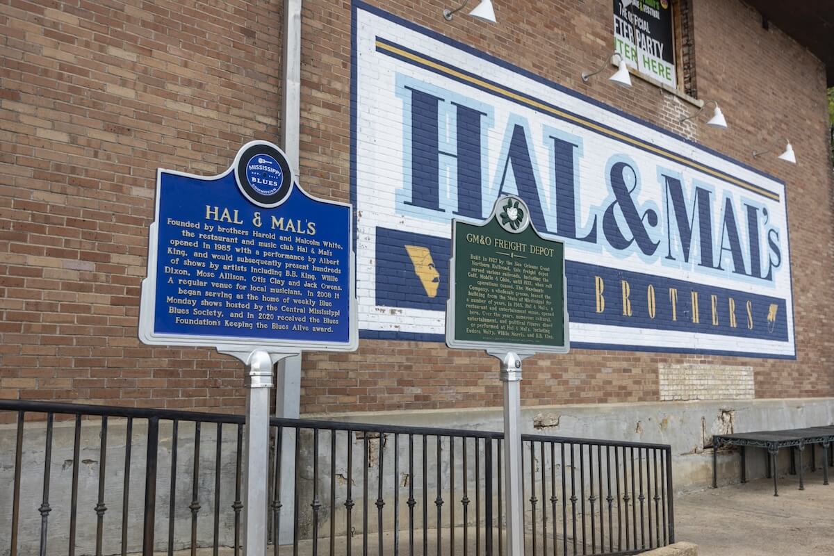 Historic markers grace the front of a brick building with "Hal & Mal's" painted on the wall, while metal railings border a walkway that hints at the South's charm.