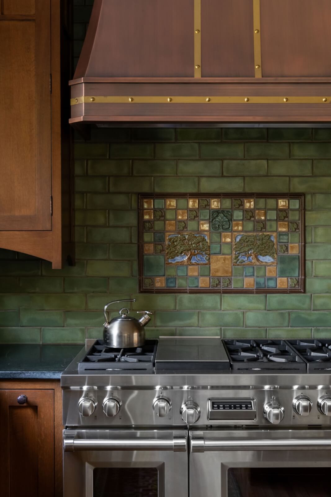 2 Homes by Beth Haley Design: A Study in Style & Contrast - 16 A stainless steel stove with four burners and a kettle is set against a green tiled backsplash featuring a tree mosaic. A copper hood and wooden cabinets surround it, blending old and new elements seamlessly in this stunning home design by Beth Haley Design.
