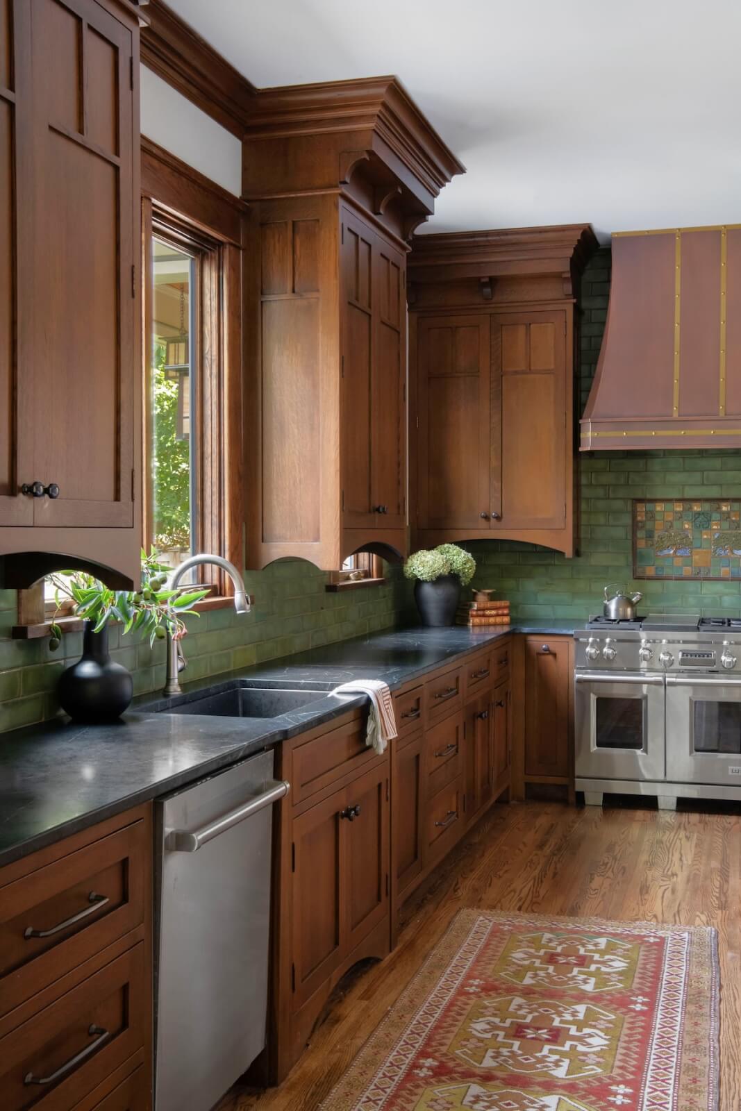 2 Homes by Beth Haley Design: A Study in Style & Contrast - 15 This stunning kitchen by Beth Haley Design beautifully marries old & new with wooden cabinetry, dark countertops, and a green tile backsplash. Stainless steel appliances complement the modern touch, while flowers by the sink and a patterned rug add warmth to this inviting space.