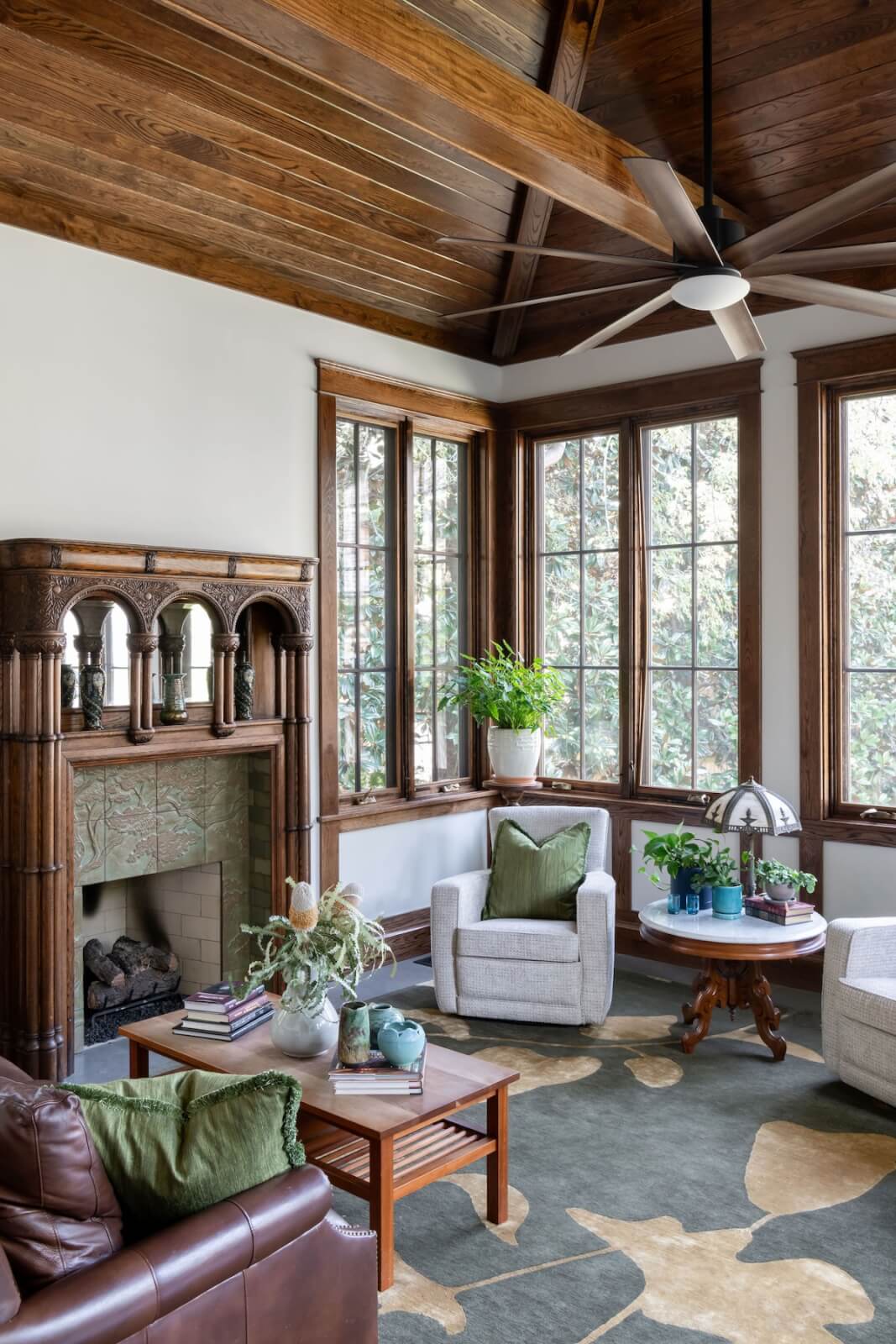 2 Homes by Beth Haley Design: A Study in Style & Contrast - 12 This stunning living room designed by Beth Haley Design features a wooden ceiling, large windows, and a cozy fireplace. Two armchairs, a leather sofa, and a round table adorned with plants rest on a patterned rug, creating the perfect blend of comfort and style.