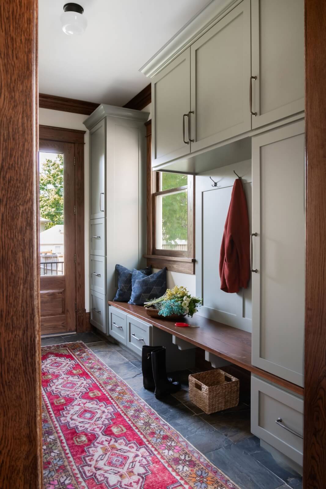 2 Homes by Beth Haley Design: A Study in Style & Contrast - 13 This mudroom, designed by Beth Haley Design, blends old and new with its green cabinets, wooden bench, and red patterned rug. A basket, black boots, cushions, and a red jacket add personality, while a bouquet of flowers graces the bench in this stunning home vignette.