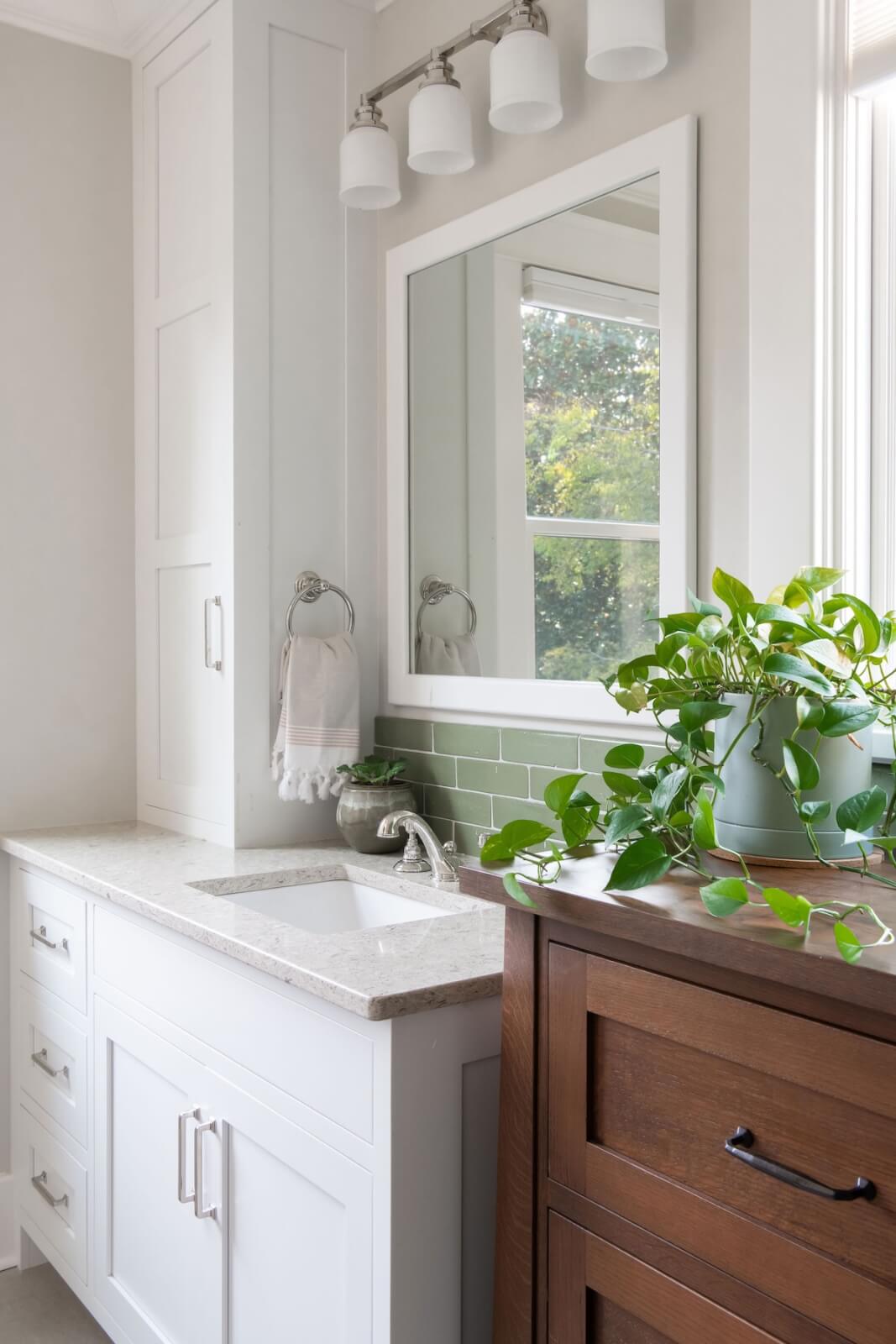 2 Homes by Beth Haley Design: A Study in Style & Contrast - 14 This stunning bathroom by Beth Haley Design blends old and new perfectly, featuring a white vanity, mirror, and countertop. A potted green plant complements the wooden cabinet. The wall's green tiles and the light fixture with three bulbs above the mirror add a touch of elegance.