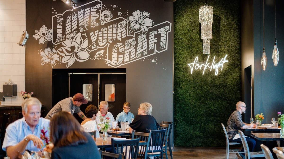 People dine in a modern Tupelo MS restaurant with black walls, graffiti art reading "LOVE HAND CRAFT," and a neon "forklift" sign on a green moss wall—perfect for trip planning or your next girls' trip.