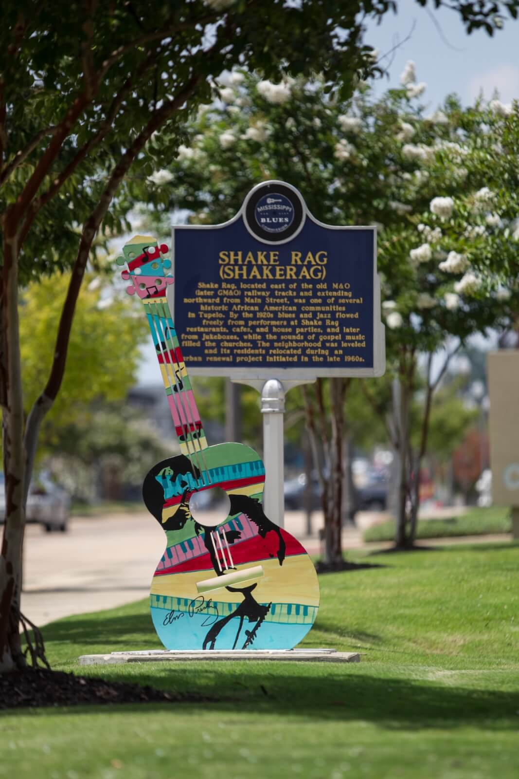 A colorful painted guitar sculpture stands on grass in front of a historical marker sign for Shake Rag in Tupelo, MS—perfect for a girls' trip photo stop beneath the shade of tree-lined streets.