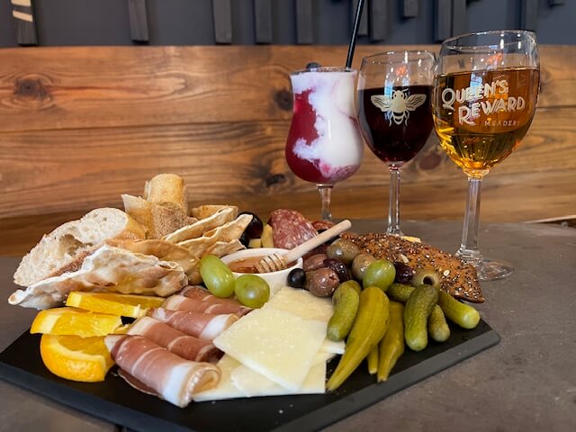 A charcuterie board with cheeses, meats, olives, pickles, and bread sits in front of three glasses of drinks—a perfect spread for a girls' trip or travel planning session in Tupelo MS.