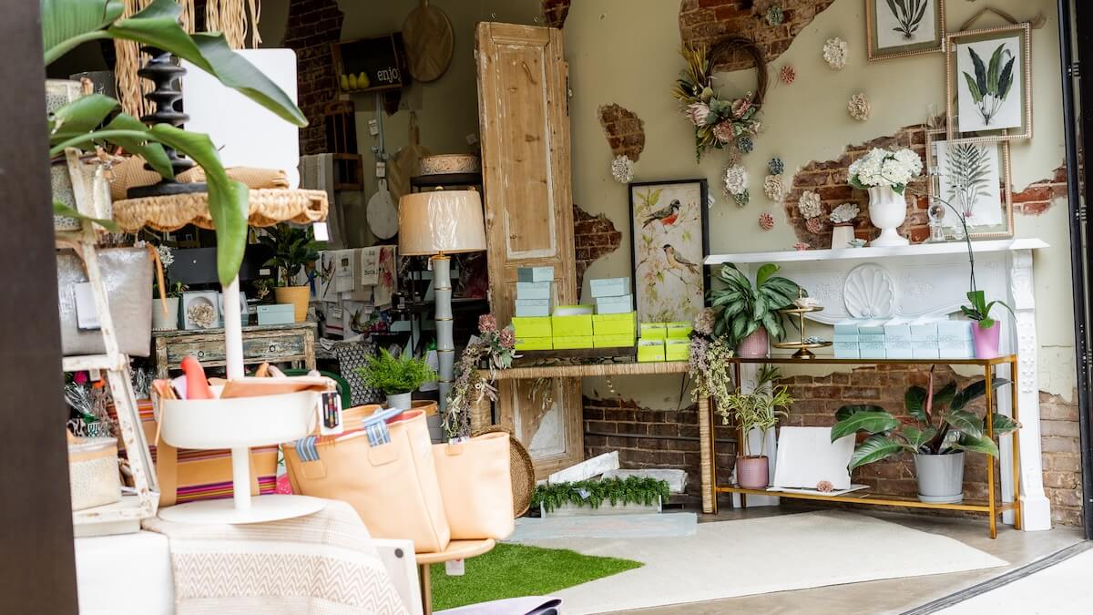 A boutique shop interior in Tupelo MS travel style, displaying plants, floral decor, handbags, pastel boxes, and framed botanical prints on a partially exposed brick wall—perfect inspiration to plan a girls' getaway.