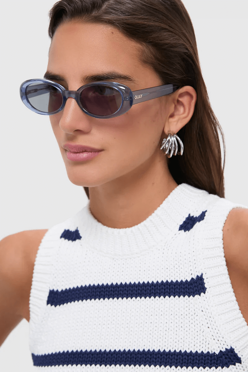 A stylish woman wears oval blue sunglasses, silver hoop earrings, and a white knit sleeveless top with navy blue stripes—perfect for summer.